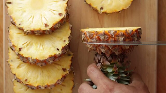 CU Close-up of Man's hand slicing pineapple with a knife on a wooden cutting board.
