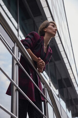 young woman with short hair in red checkered jacket outdoors in front of office building. portrait of caucasian female leaning on railings at street
