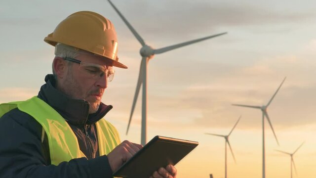 Renewable energy technician engineer using tablet,wind turbine plant in the background,worker or operator checking wind turbine does mainteinance to the power station generator