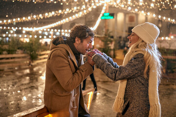 A young guy is warming up the hands of his girl while they walking under christmas lights in the city. Christmas, New Year, holiday, love