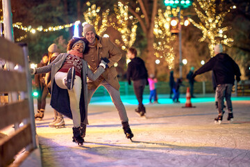 A young guy is helping his girlfriend while learning to skate at ice-skating rink during christmas holidays in the city. Christmas, New Year, holiday, love