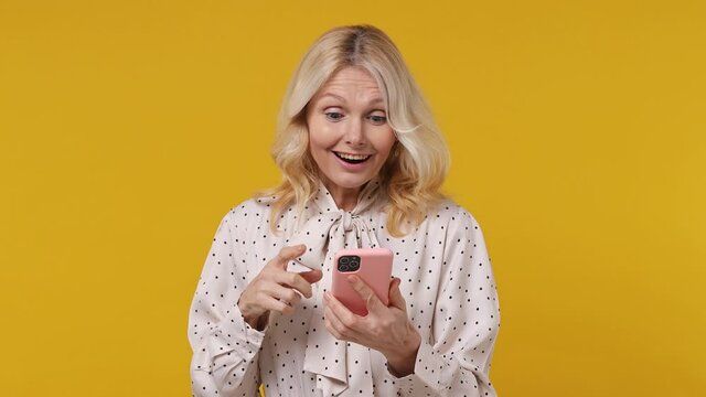 Happy Excited Elderly Blonde Woman Lady 40s Years Old In Pink Shirt Hold Use Mobile Cell Phone Typing Say Wow Yes Just Found Out Great Big Win News Isolated On Plain Yellow Background Studio Portrait