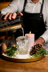 Bartender pouring strong and fresh drink into the cocktail glass on the plate decorated with rosemary branches, pine cones and lighting candle.