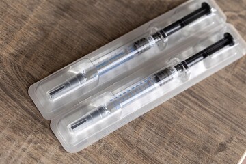 A portrait of a package with two syringes in it lying on a wooden table. The drug injection shots are made of a glass tube with marks volume marks on it and the needle still has the cap on it.
