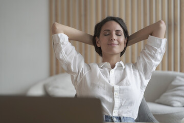 Positive female entrepreneur sitting at desk at home in relaxing position with hands behind her head