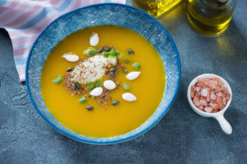 Pumpkin soup topped with soft cheese, pumpkin seeds and scallions, elevated view on a blue stone background, horizontal shot