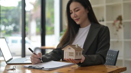 Photo of a beautiful Asian woman writing on paperwork and holding a house model while sitting at the wooden working desk.