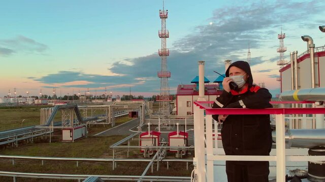 An oil and gas industry worker, a man wearing a mask and dripping clothes and overalls, stands on a tower near a rectification tower at an oil refinery. The mechanic speaks on a walkie-talkie.