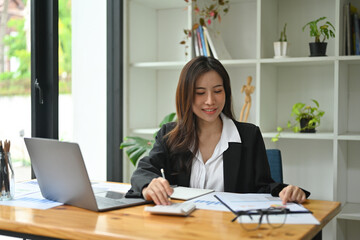 Photo of a beautiful insurance agent using a computer laptop at the wooden working desk surrounded by a document and office equipment.