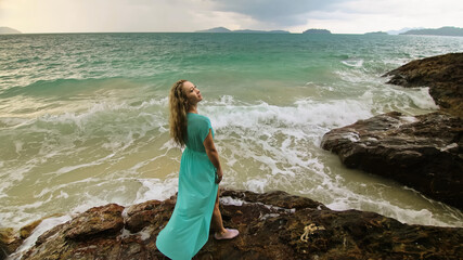 Woman walks on rock of sea reef stone, stormy cloudy ocean. Blue swimsuit dress tunic. Concept rest, tropical resort coastline tourism summer holidays