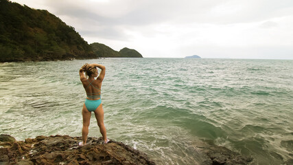 Woman chill relax resting on rock of sea reef stone, stormy cloudy ocean. Woman in swimsuit dress tunic. Concept rest tropical outdoor tourism