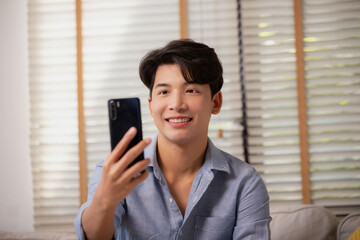 Fototapeta premium Asian man making facetime video calling with smartphone at home. He's waving on phone screen. Using conferencing meeting online app.