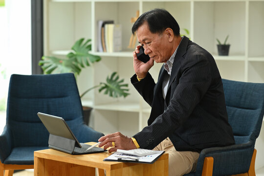 Photo of an elderly businessman talking on mobile phone while using a digital tablet and stylus pen at the comfortable chair.