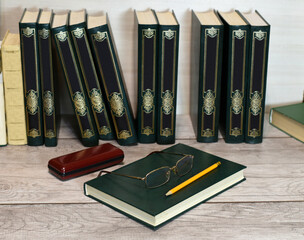 There are several books on the shelf with the same beautiful binding. Nearby lies a book with glasses and a pencil, nearby is a case for glasses.