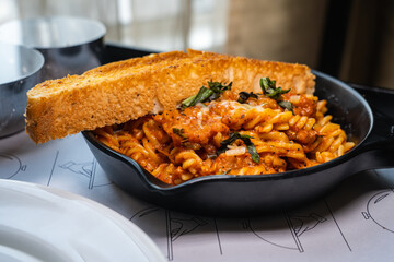 Fusilli Arrabiata pasta and a slice of bread served on black iron pan.