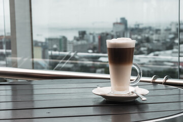 A glass of coffee latte on a table against a blurred background of a cityscape.