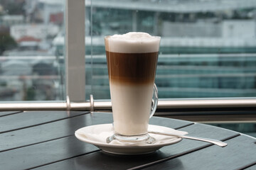 A glass of coffee latte on a table against a blurred background of a cityscape.