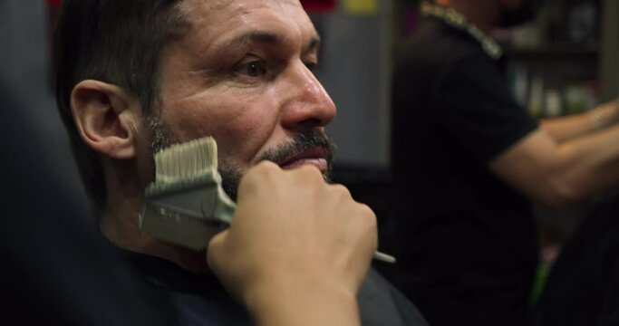 Haircut beard with in a barber shop. Handsome man in a barbershop shaves his beard in a hair studio. A professional hairdresser paints a client man beard with black hair dye.