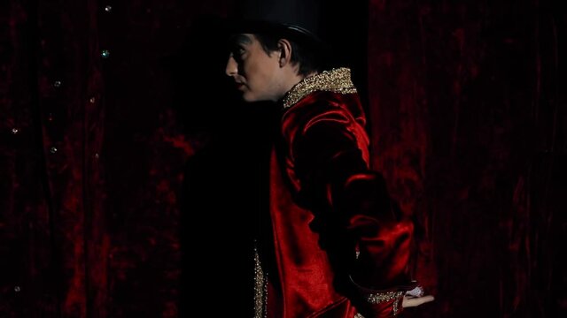 The showman effectively begins to show the trick with his fingers, passes through the curtain, enters the stage. The guy is a wizard with makeup in a red dress coat and top hat. A bright suit against 