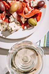 cool white wine in a glass near a plate with sliced jamon, dove cheese and various olives. a light snack on a hot day. classic italian food.