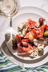 cool white wine in a glass near a plate with sliced jamon, dove cheese and various olives. a light snack on a hot day. classic italian food.