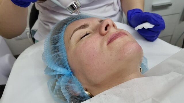 Beauty salon. A cosmetologist in medical gloves and protective mask doing a hydra peeling procedure on the client's cheeks. Side view. Close up. Professional skin care during coronavirus pandemic.