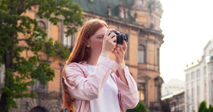 Pretty female lady walking on street in city, taking pictures and looking at antique buildings. Redheaded girl photographer using digital camera. Technology, tourism, traveling concept.
