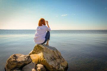 A young girl in a white shirt sits barefoot on the rocks near the shore and looks into the distance. A beautiful, slender woman is resting on the sea coast.