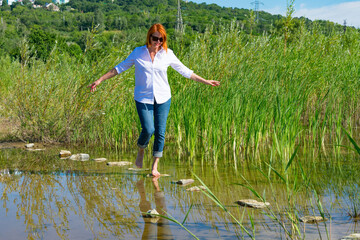  A young woman in a white shirt runs barefoot down the river. The woman crosses the stream over the...