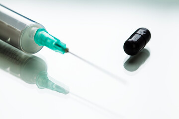 Green needle in syringe and black pill medication on white reflective surface