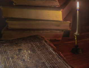 Candle in candlestick illuminates cover of old bible and books in the background