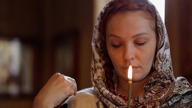 a Russian woman in a headscarf holds a candle in her hands and stands in front of icons in an Orthodox church and prays