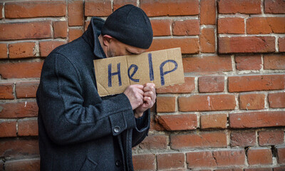 A man in an old coat stands at a brick wall with a sign in his hands and asks for help. Social themes, poverty and vagrancy.