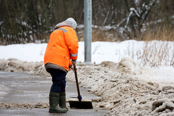 Snow removal in winter city, woman worker in uniform with a shovel cleaning the street near the park