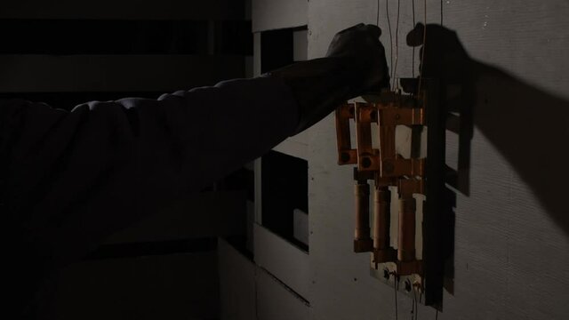 In dramatic lighting a black rubber gloved hand reaches into the frame to grab the wooden handle of an old large industrial electrical bar switch to pull it open (off) then withdraws from the scene. 