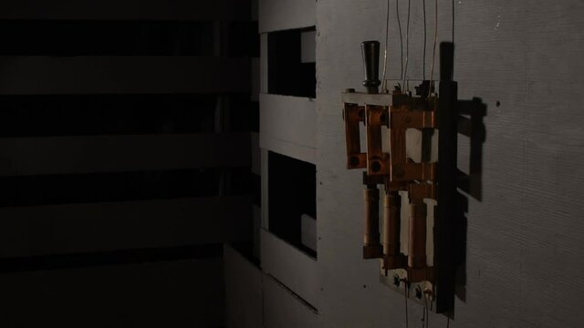 In dramatic lighting a black rubber gloved hand reaches into the frame to grab the wooden handle of a close old large industrial electrical bar switch, gives it a little shove to be sure it is on. 
