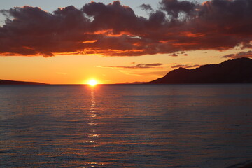 Sunset on the sea, silhouette of mountains against the background of an orange sky with picturesque clouds and the sun on the horizon