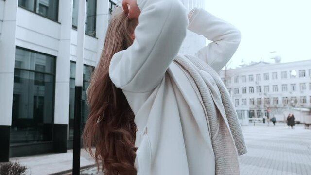 A girl in a white coat and a scarf straightens her thick hair. Strong wind. Business center in the background. Success