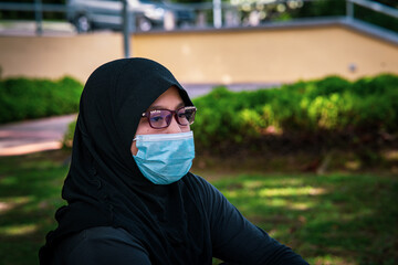 Portrait of young muslim woman wearing glasses and hijab head scarf and face mask. Casual islamic girl at the outdoor park.