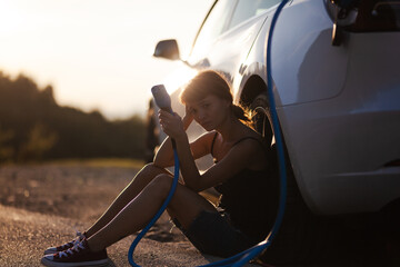 Beautiful young sad girl next to an electric car. Holding a charging cable. Sunset backlight.