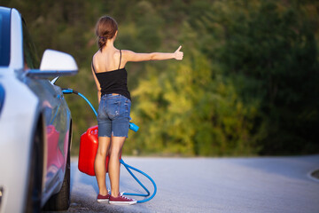Girl standing next to electric car holding a charging cable and gassoline canister while hitchhiking