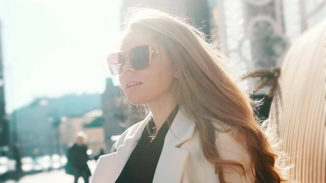 A portrait of a girl with glasses in the bright sun straightens her gorgeous blond hair with her hand, a busy city in the background. Close-up, fashion