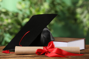 Graduation hat, book and diploma on wooden table against blurred background