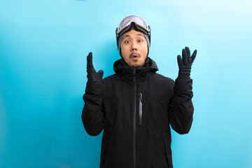 Handsome asian man with snowboard goggles and black anorak coat surprise expression on blue...