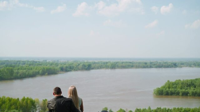 Lovely couple of blonde lady and motorcyclist man sits resting on motorcycle on green hill near tranquil river on spring day backside view slow motion