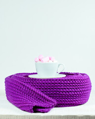 Hand-knitted scarf and cup with marshmallows on white table Velvet violet