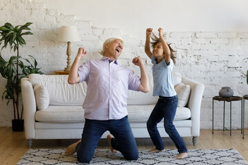 Overjoyed mature senior retired grandfather standing on knees, dancing to energetic disco rock pop music with emotional laughing preteen grandchild boy, having fun enjoying weekend activity at home.