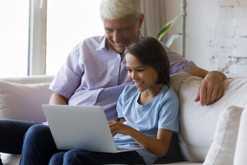 Happy small adorable preteen schoolboy using computer apps with joyful caring middle aged mature retired grandfather, sitting on couch at home, playing online games, shopping in internet store.