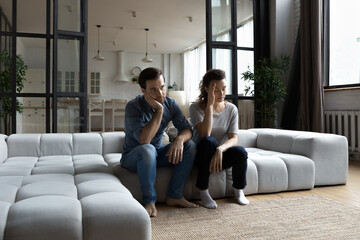 Tired unhappy young couple sitting on couch at home, thinking about relationship problems after...