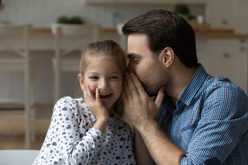 Excited little girl and father sharing secrets or funny news, loving Caucasian dad whispering in adorable 8s girl child ear, family having fun together, talking, enjoying leisure time at home